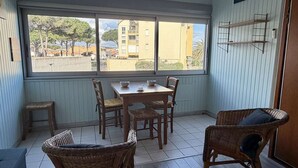 Uma área de refeições com uma mesa e cadeiras, uma janela com vista para prédios e uma estante com objetos.