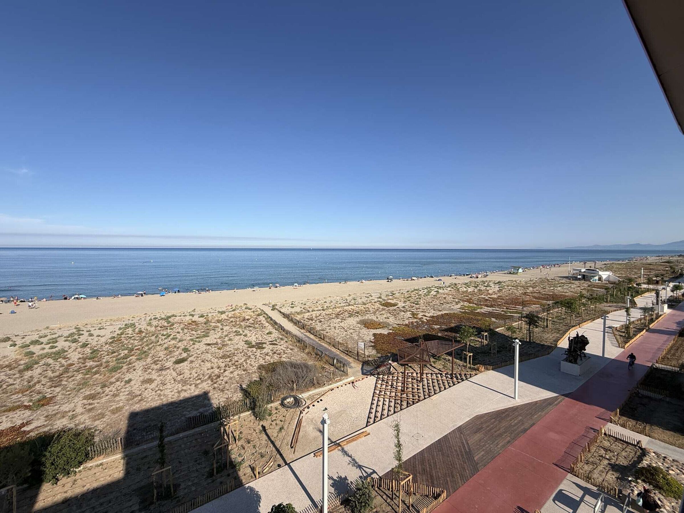 A beach view with a clear sky, a sandy beach, and a paved walkway leading to a building.