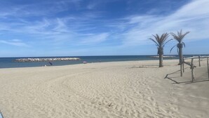 A sandy beach with palm trees, a clear sky, and a rocky breakwater in the distance. - Duplex with sea view, AC - 6 guests (Le Barcarès)