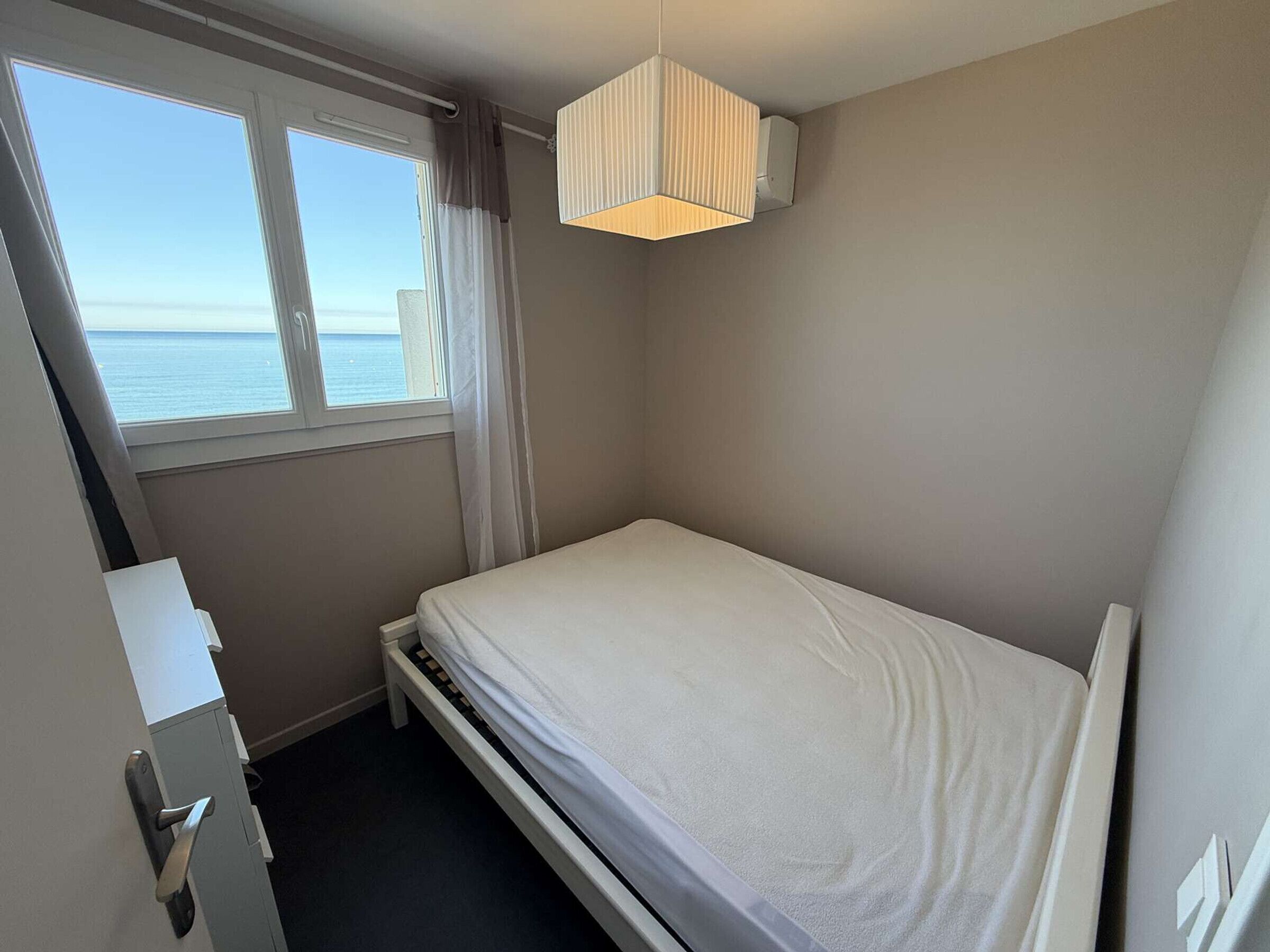Pequeña habitación con cama individual, ventana con vista al mar, mesita de noche y lámpara colgante.