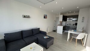 A modern living room with a dark sofa, a white coffee table, and a kitchen in the background. - Modern apartment, dune view - 2 guests (Saint-Hilaire-de-Riez)
