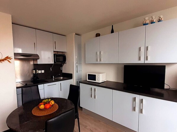 A compact kitchen with white cabinets, a microwave, a black countertop, and a dining table with fruit. - Charming duplex lake view - 4 guests (Le Barcarès)
