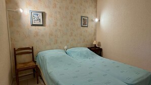 A small room with a bed, a chair, two framed pictures on the wall, and a bedside table with a lamp. - Apartment with port view - 4 guests (Gruissan)
