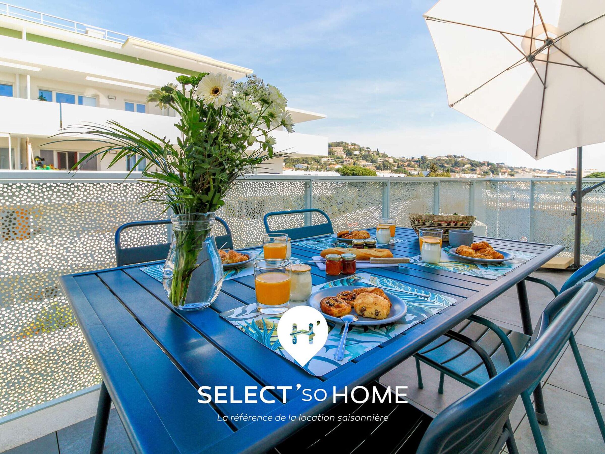 A dining table set with breakfast items, a vase of flowers, and a view of a coastal town.