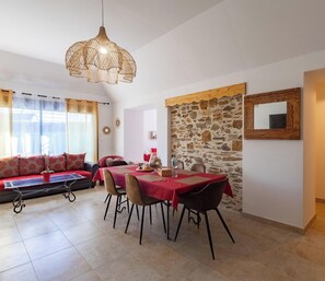 A dining area with a table set for four, a sofa with cushions, a hanging light fixture, and a stone wall. - Cozy cottage near Angers - 5 guests (Chambellay)