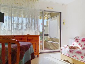 Ein Zimmer mit einem Holzstuhl, einem Tisch mit Tischdecke, einer Couch mit gemusterten Kissen, einer Schiebeglastür und Blick auf ein Gebäude.