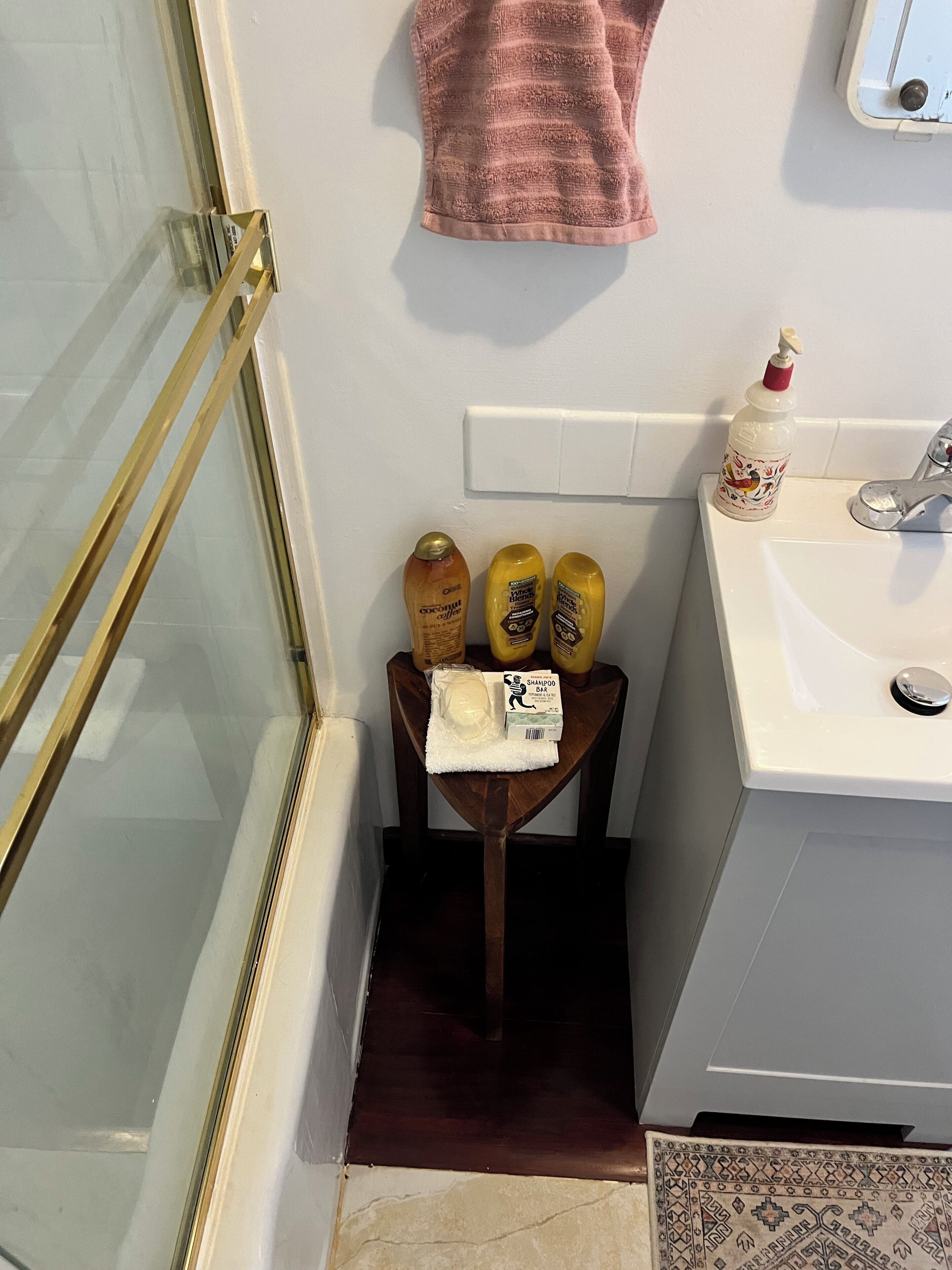 Casa de banho com chuveiro em vidro, uma cadeira de madeira, pia branca e produtos de higiene numa prateleira.