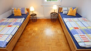 A room with two wooden beds, each with a colorful striped bedspread and a yellow pillow. There are two round, wireframe nightstands with lamps between the beds. A window with a white radiator is above the beds. - Apartment 35qm (Villingen-Schwenningen)