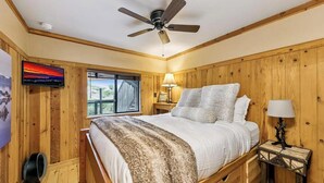 Een slaapkamer met een bed, een plafondventilator, een nachtkastje met een lamp, een raam met uitzicht en een aan de muur bevestigde televisie.