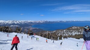 Een skipiste met skiërs, een helderblauwe lucht en een bergketen op de achtergrond.