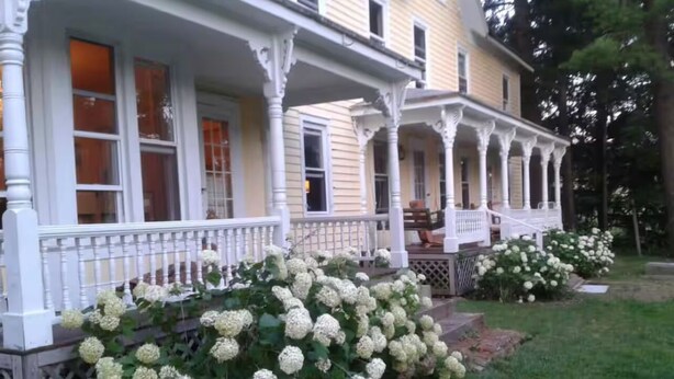 A two-story house with a porch and a garden with hydrangea bushes. - Near The Baseball Hall of Fame, Glimmerglass Opera and Dream Park Baseball Camp (Cherry Valley)