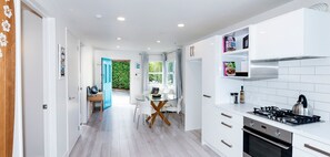 A modern kitchen with white cabinets, a gas stove, and an oven. There is a dining area with a glass table and chairs. - Welcome to nature and waves in Auckland perfect for your getaway (Auckland)