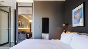 Une chambre d’hôtel avec un grand lit, un bureau avec une chaise, un tableau encadré au mur et une porte donnant sur une autre pièce.