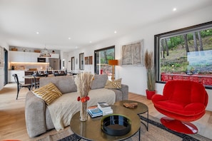 Ein modernes Wohnzimmer mit einer Couch, einem roten Sessel, einem Couchtisch und einem Essbereich, im Hintergrund eine Küche.