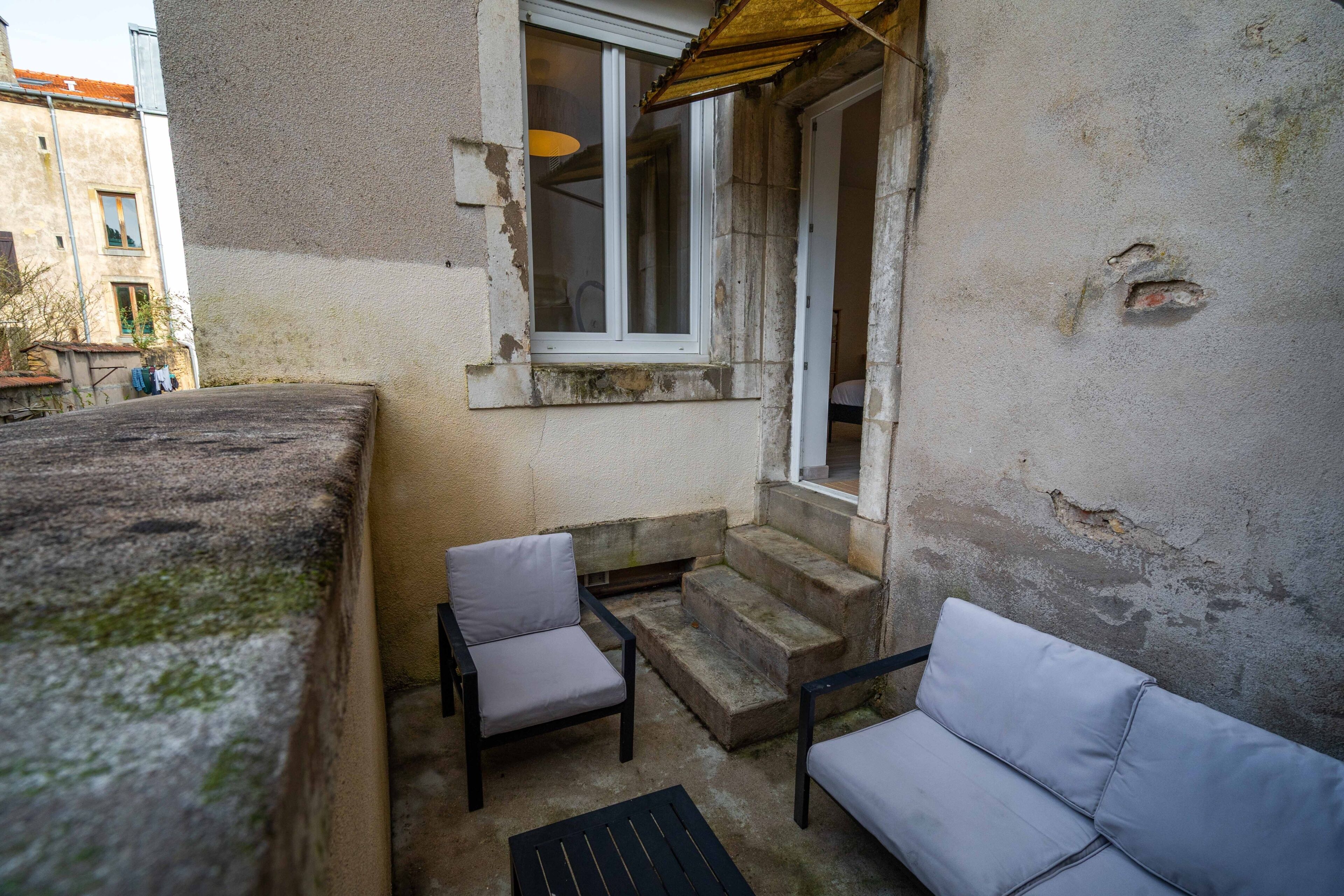 En lille udendørs siddeplads med en sofa og to stole, et vindue med udsigt og en trappe af sten, der fører til en dør.