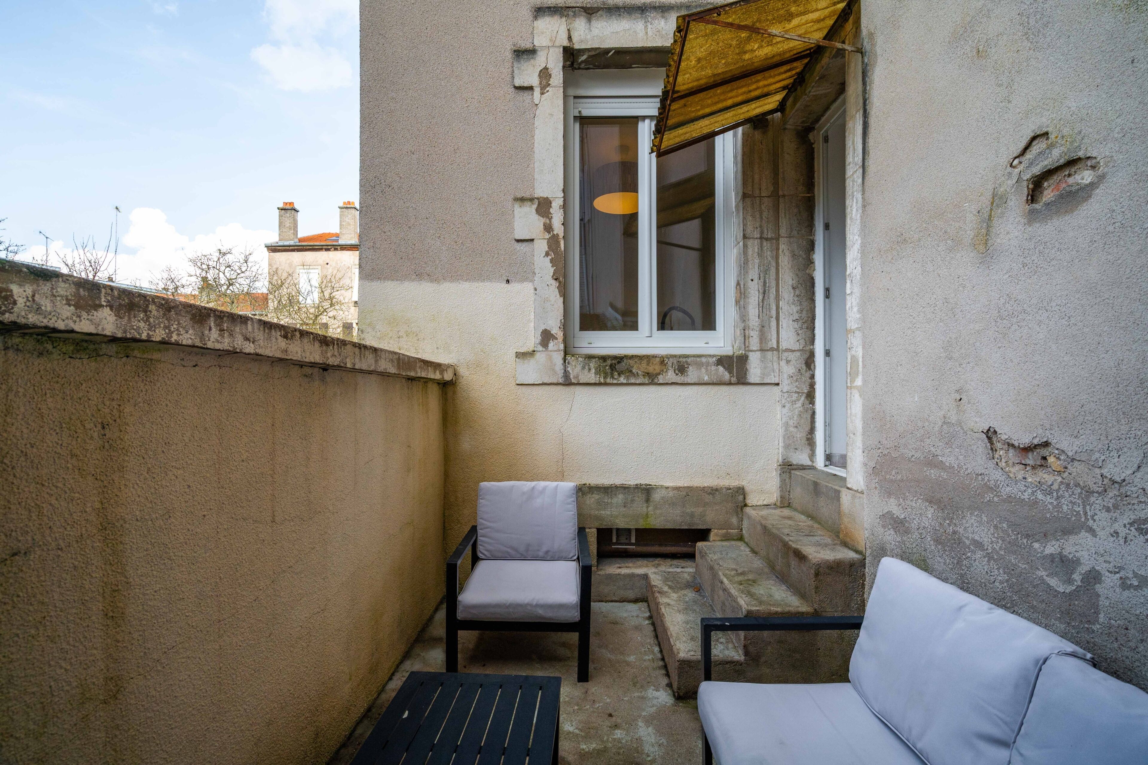 Ein kleiner Balkon mit Betonwand, einem Fenster mit gelbem Markisenvorzelt und zwei Stühlen.