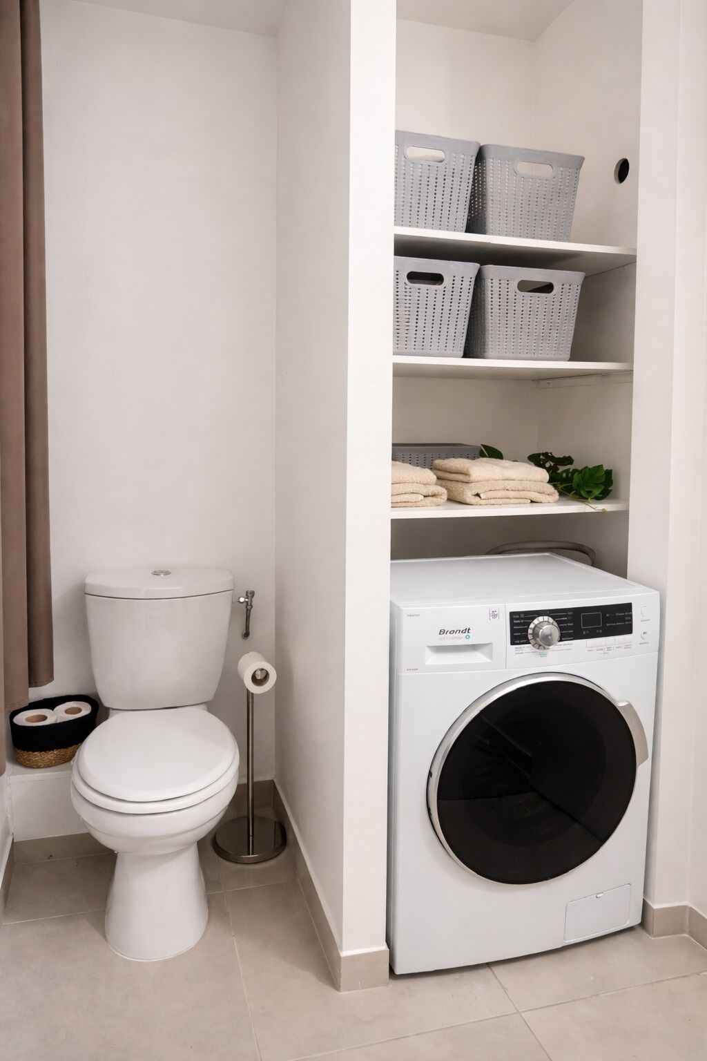 A compact bathroom with a toilet, a washing machine, and shelves with baskets.