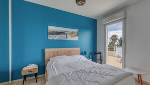 A bedroom with a bed, a wooden nightstand, a small table, a painting on the wall, and a view through an open door. - La Terrasse de La Ciotat - climatisé, proche plage (La Ciotat)
