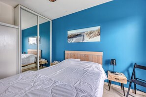 Un dormitorio con cama, mesita de noche, silla, lámpara y un cuadro en la pared.
