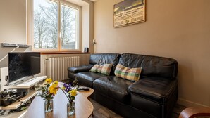 Ein Wohnzimmer mit einer schwarzen Ledersofa, einem Couchtisch mit Blumen, einem Fernseher und einem Fenster mit Blick auf Bäume.