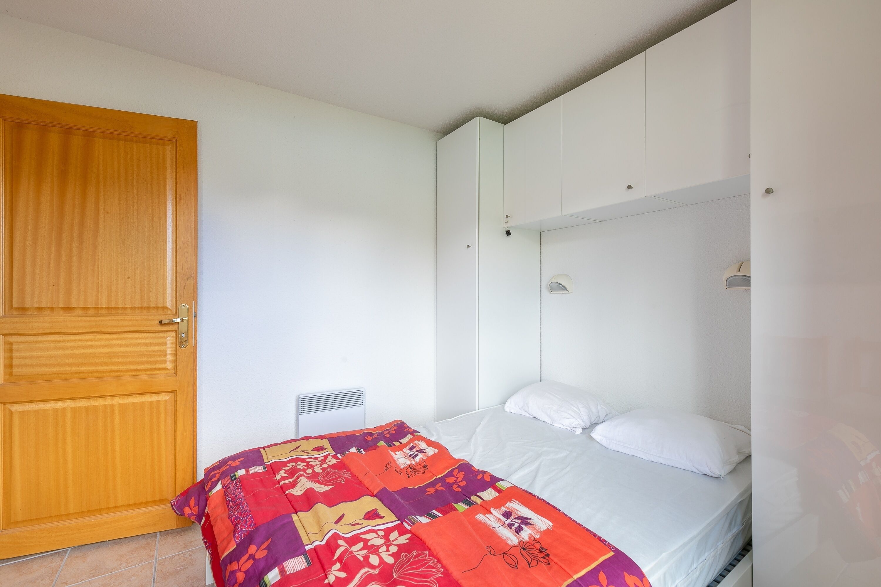 Un dormitorio con puerta de madera, cama con manta colorida y paredes blancas.