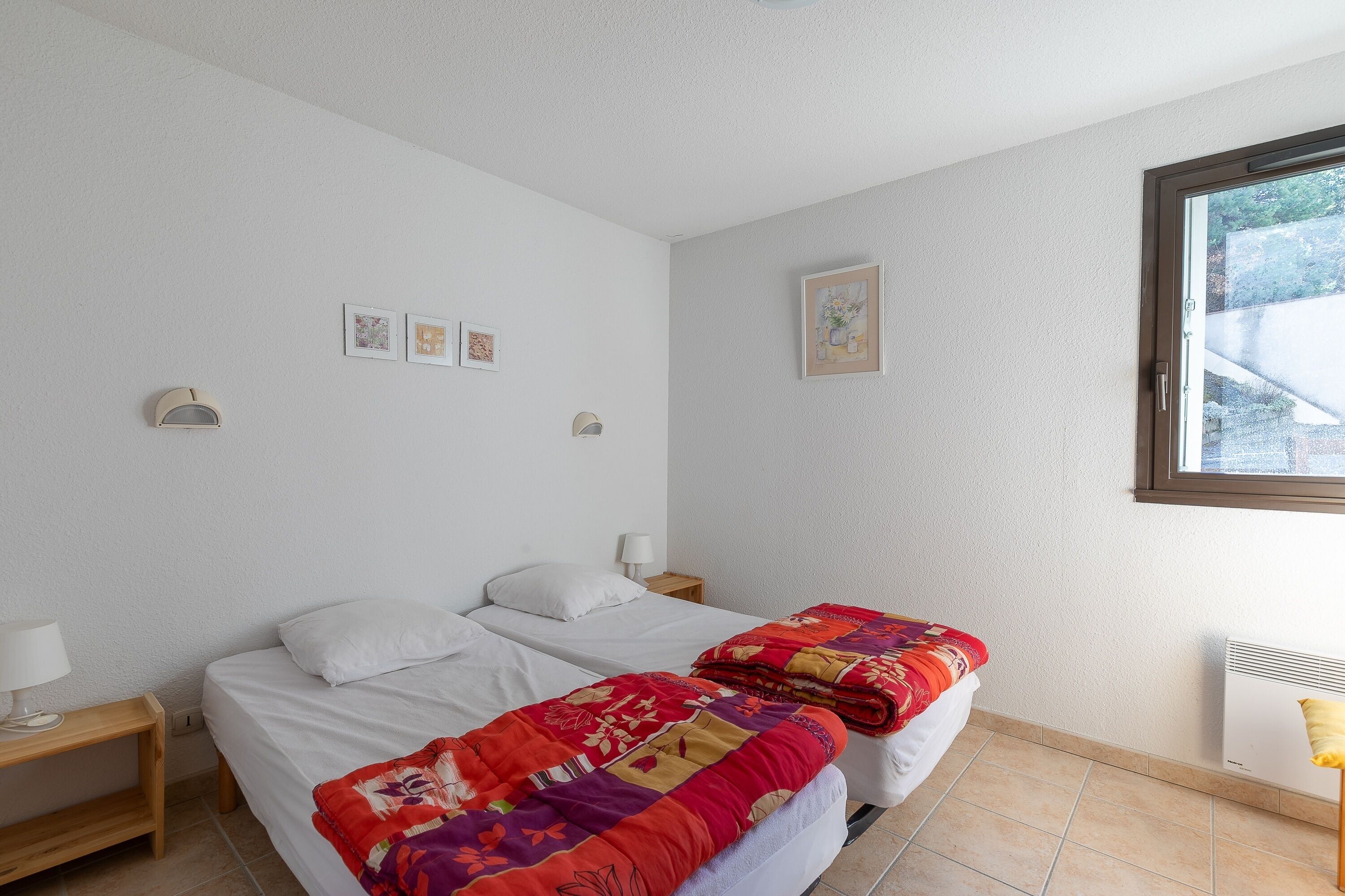 A bedroom with two beds, a nightstand, a lamp, and a window.
