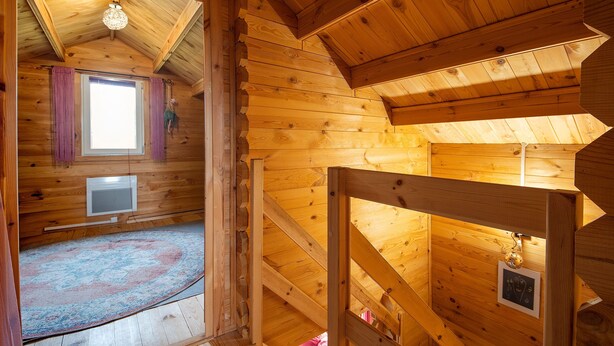 Interior de una cabaña de madera con techo inclinado, una ventana con cortinas, un televisor de pantalla plana y una escalera de madera.