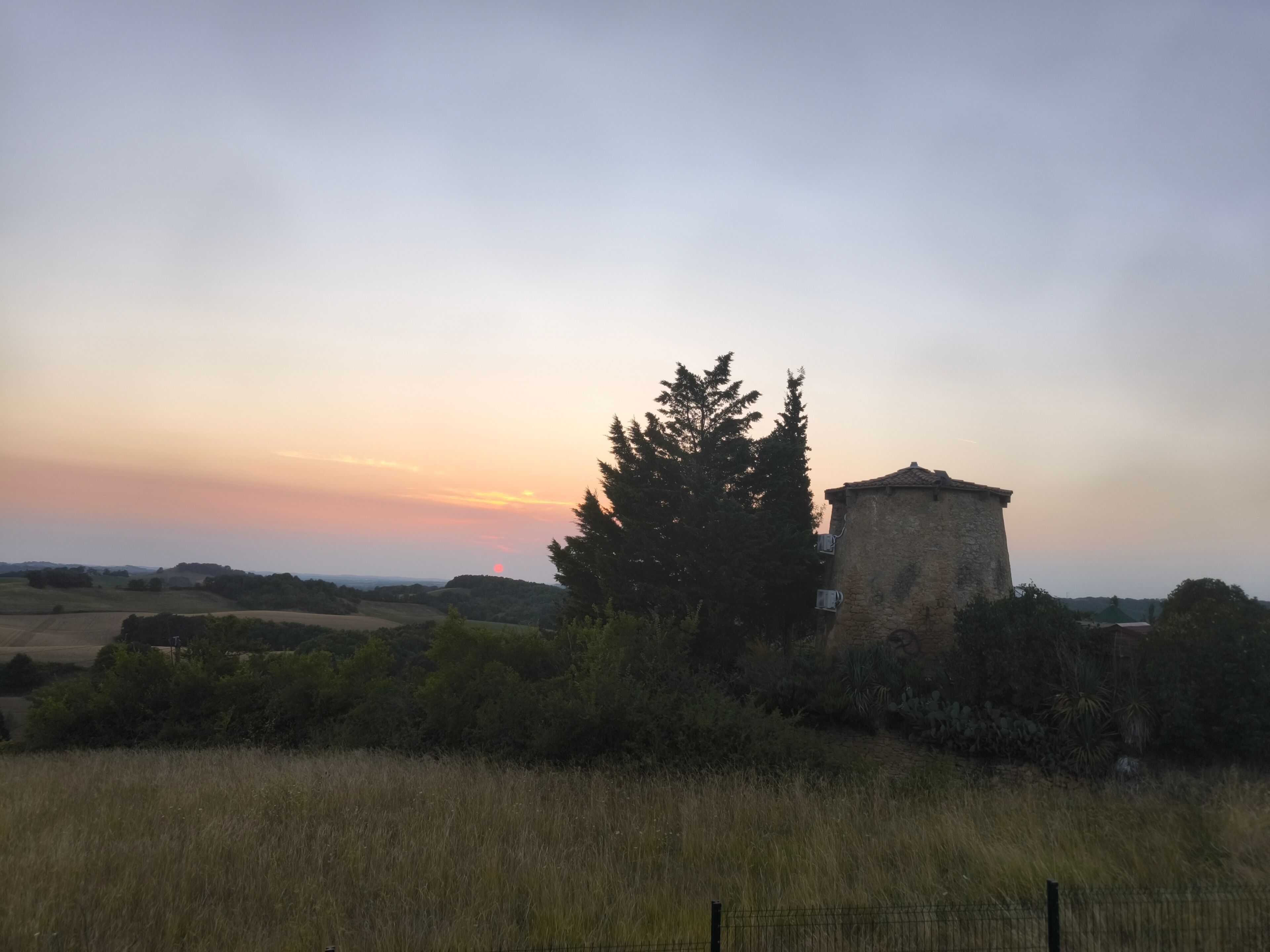 Paisaje rural con una construcción de piedra, posiblemente una torre, rodeada de campos y árboles.