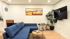 A living room with a blue sectional sofa, a round coffee table, a wall-mounted TV, a clock, and a painting.