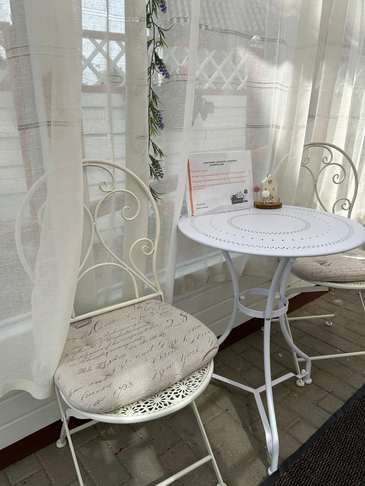 Ein weißer Metalltisch mit Stühlen und einem Kissen mit Schreibschrift, einem Schild mit Text sowie einem dekorativen Gegenstand auf der Tischfläche.