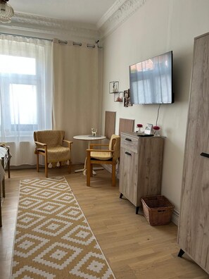 Ein Zimmer mit einem Holzkasten, einem Fernseher, zwei Stühlen, einem kleinen Tisch, einem gemusterten Teppich und einem Fenster mit Vorhängen.