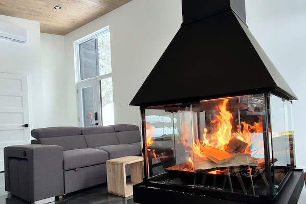 Un salon moderne avec une cheminée à façade vitrée, un canapé gris et un petit tabouret en bois.