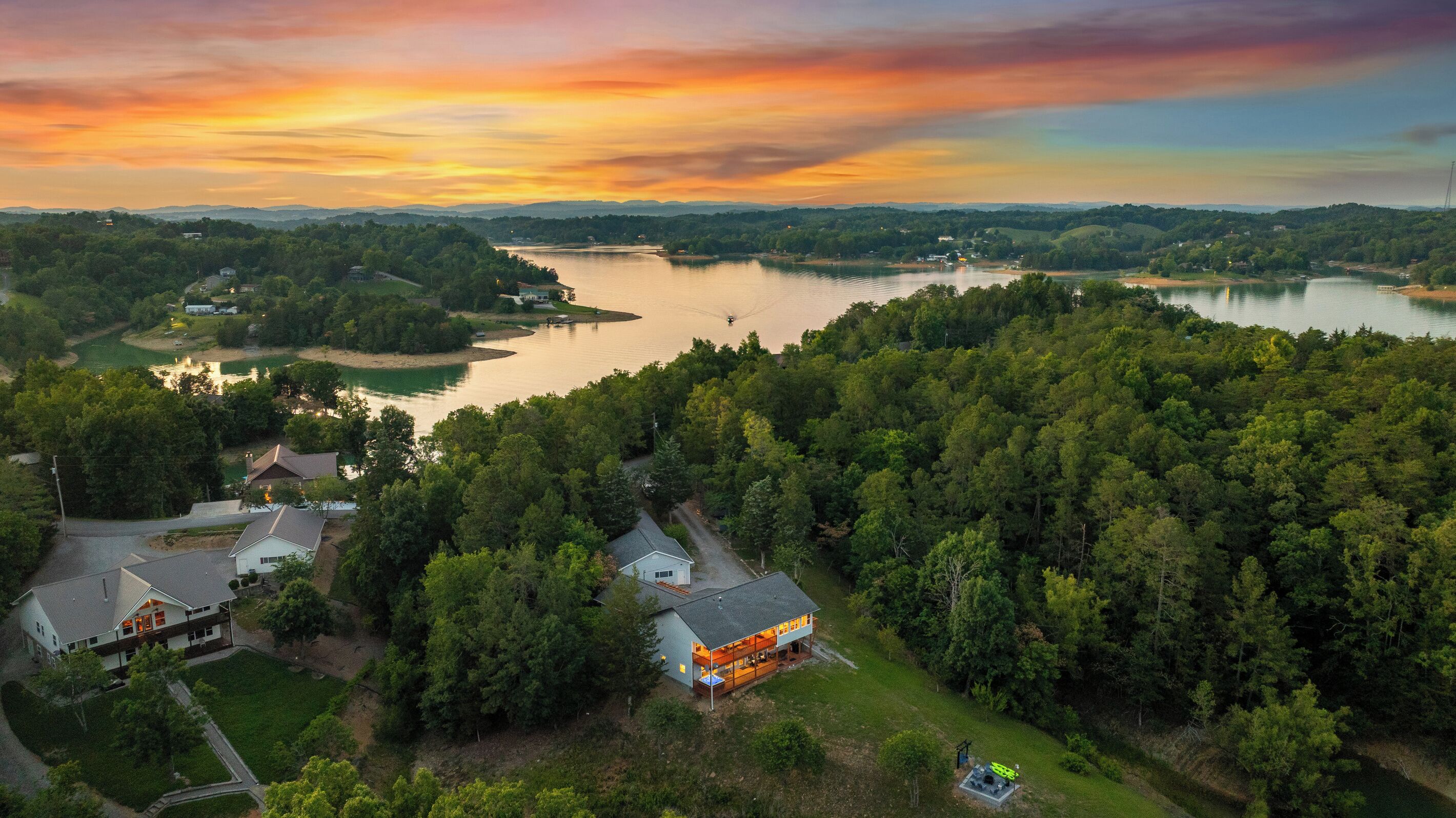 An aerial view of a residential area with houses, a lake, and a forested landscape during sunset.