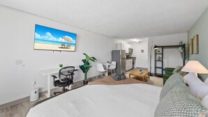 Une chambre d’hôtel équipée d’un lit, d’un bureau, d’une chaise, d’une kitchenette et ornée d’un tableau représentant une plage.