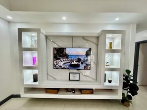 Una sala de estar moderna con un televisor de pantalla plana grande montado en una pared de mármol, rodeada de estanterías empotradas con objetos decorativos y un mueble bajo de madera.