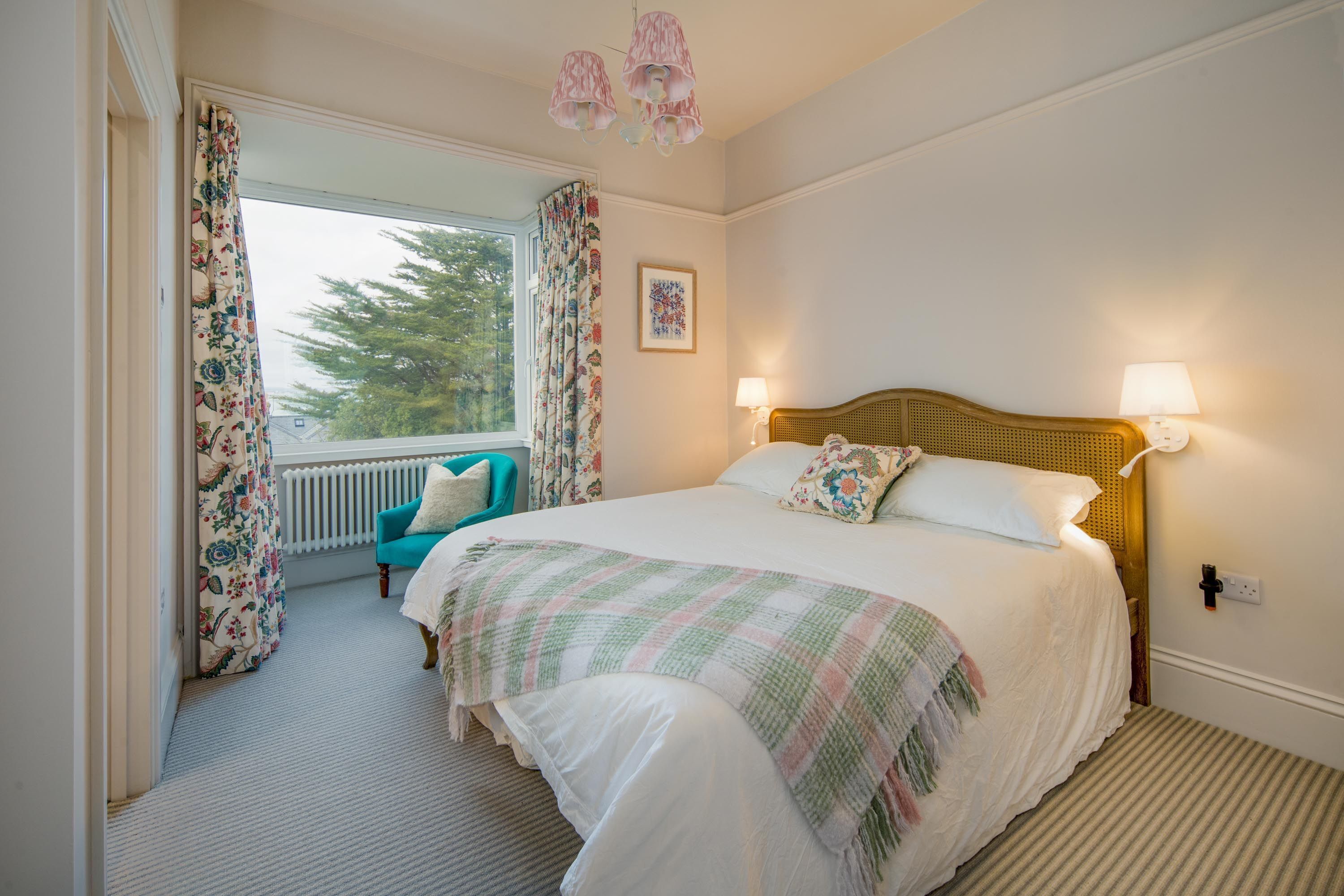 A bedroom with a bed, two bedside lamps, a window with curtains, a chair, and a radiator.
