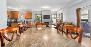 A modern kitchen with wooden cabinets, a dining area with a marble table and wooden chairs, and a living room with a sofa and a TV.