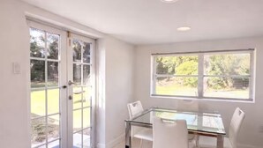 Une pièce avec une table en verre, des chaises blanches, une fenêtre avec des stores et une porte donnant sur l’extérieur.