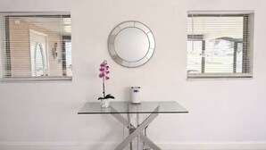 Une pièce avec une table en verre, une orchidée en pot, un miroir rond et deux fenêtres équipées de stores.