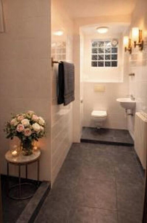 A narrow bathroom with a toilet, sink, and a small table with a vase of flowers. - City Living Trelleborg  (Trelleborg)