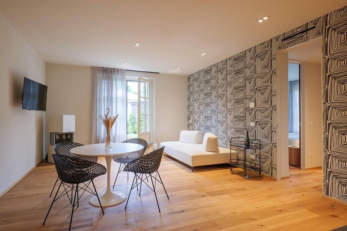 A modern living room with a round dining table, black wireframe chairs, a white sofa, and a patterned wallpaper.