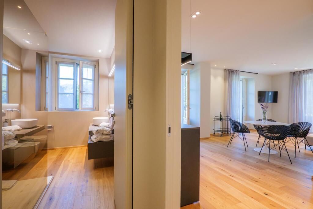 Une chambre d’hôtel moderne avec un plancher en bois, une grande fenêtre, un téléviseur à écran plat et un coin repas avec des chaises.