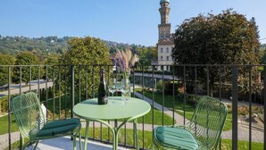 Un balcón con una mesa, sillas, una botella y copas. Al fondo, una torre y un edificio con techo verde.