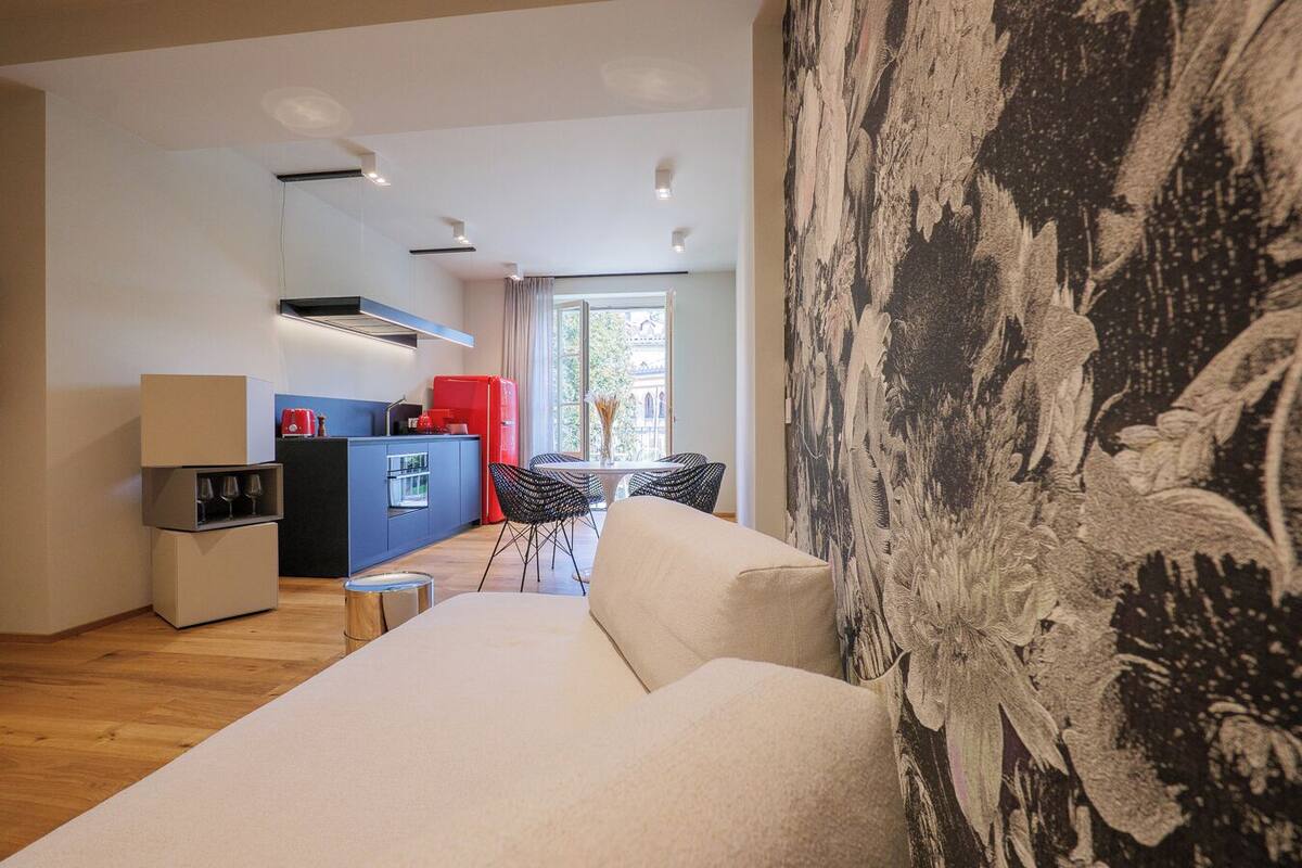 A modern living room with a sofa, a dining area with chairs, a kitchen with a red fridge, and a large window.