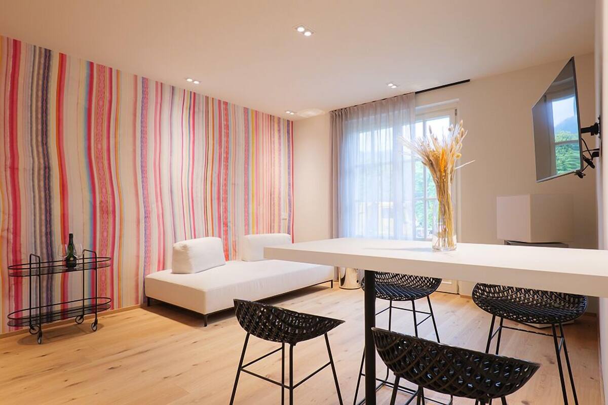 A modern living room with a white sofa, a dining table with four black chairs, and a colorful striped wall.