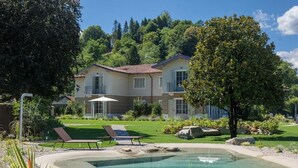 Una piscina con sillones, una casa con jardín y una colina al fondo.