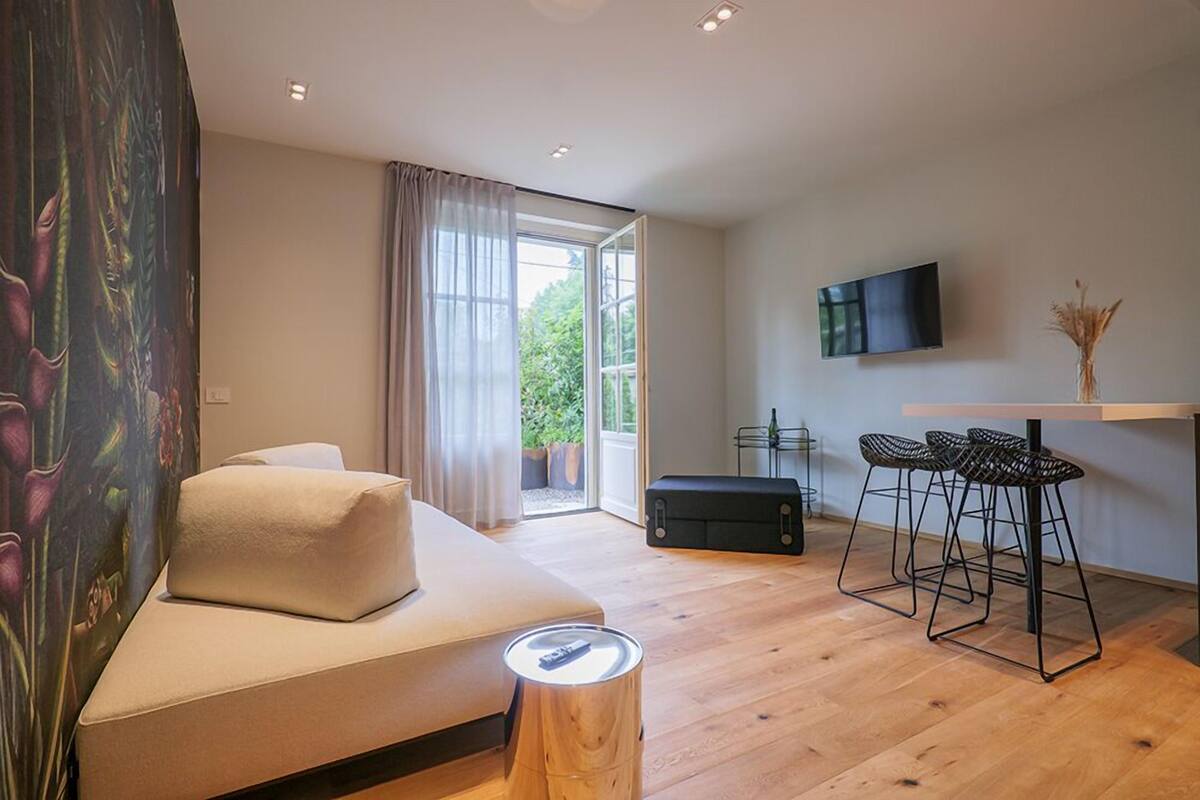 A modern room with a sofa, a TV, a dining area with stools, and a window with curtains.