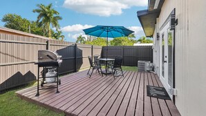 Una terraza con una parrilla, mesa y sillas exteriores, un paraguas azul y un tapete que dice 'hello'.