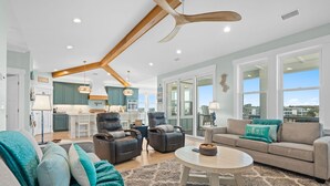 A spacious living room with a large ceiling fan, a dining area, and a kitchen with teal cabinets. - Into The Mistic, Ocean Isle Beach (Ocean Isle Beach)