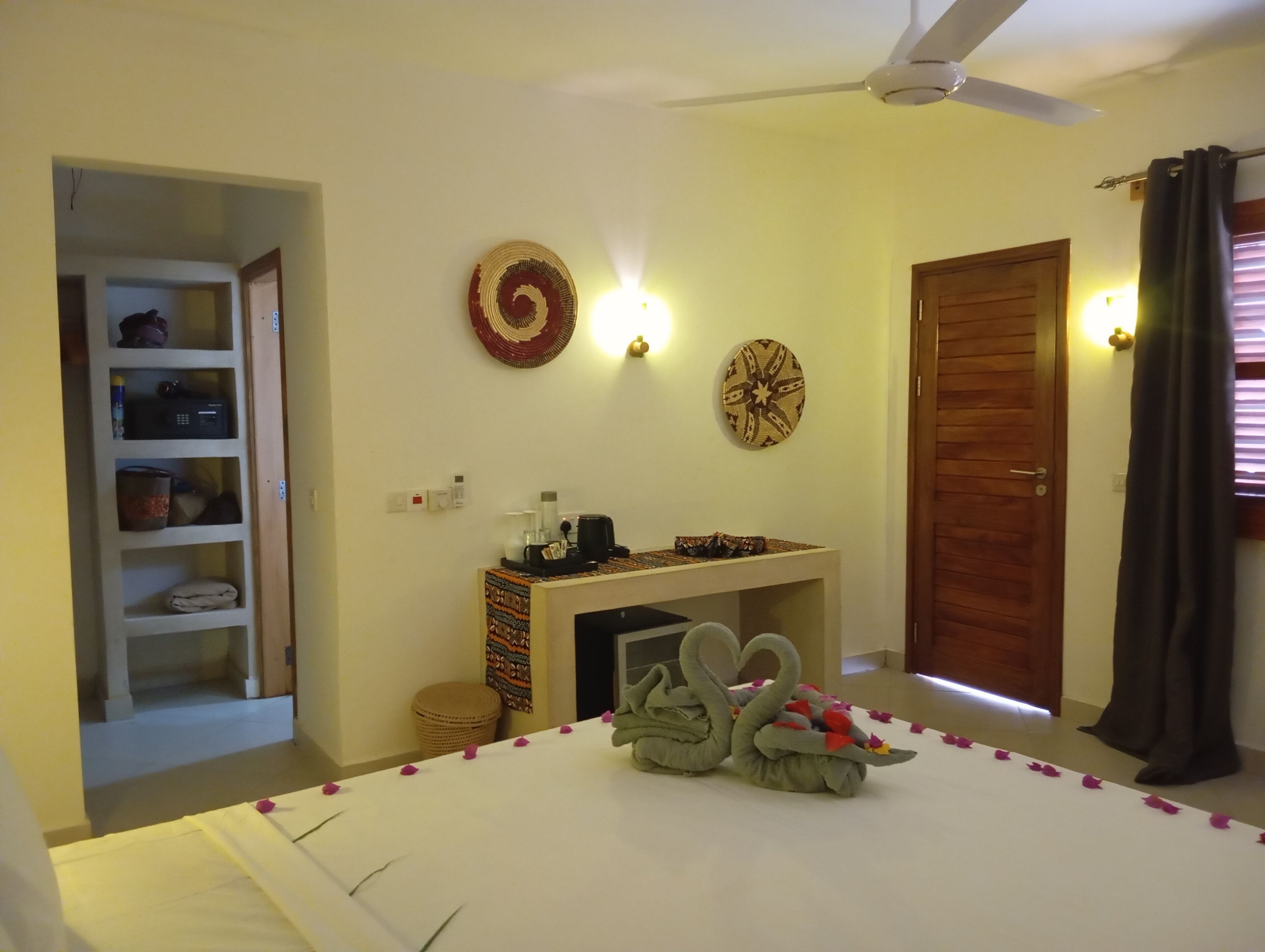 A bedroom with a bed, a ceiling fan, a small table with a decorative item, a wall with two wall-mounted lights, a door, and a shelf with various items.